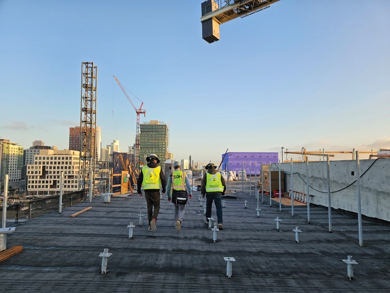 Three construction workers in safety vests and helmets walk across a rooftop under construction with a clear sky and cityscape in the background.