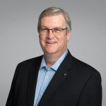 Smiling man in glasses with grey hair wearing a dark suit jacket and light blue shirt. Neutral gray background. Professional and approachable tone.