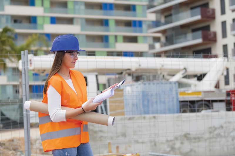 A woman in a hard hat and safety vest, holding blueprints and documents, stands at a construction site. Modern buildings are visible in the background.