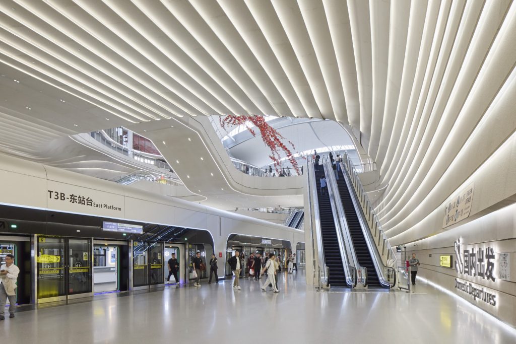 Futuristic airport interior with curved, ribbed walls, escalators leading up, and people walking. Sleek, modern design with soft lighting.