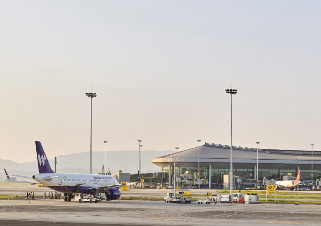 A purple and white airplane is parked on a sunny airport tarmac near a modern terminal building. Ground vehicles and personnel are nearby, with mountains in the background.