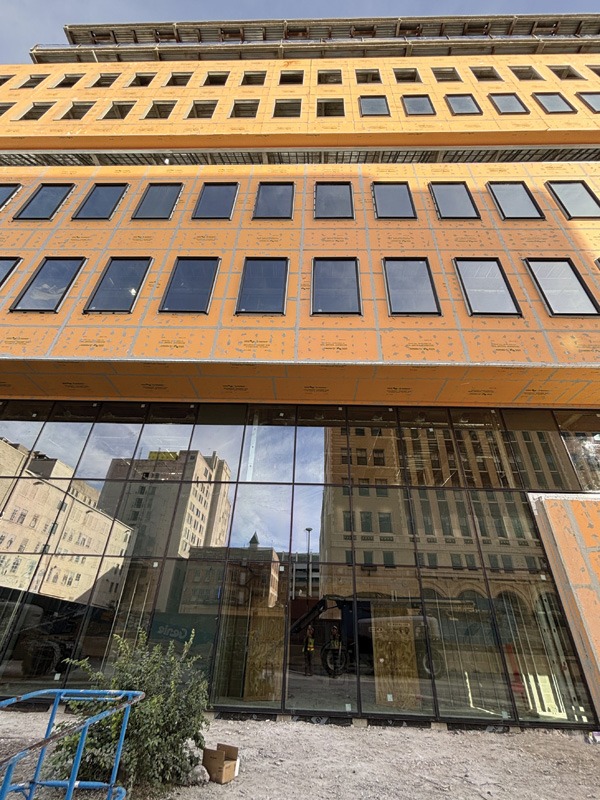 A partially constructed building with orange sheathing, featuring large reflective glass windows. The urban setting is visible in their reflection.