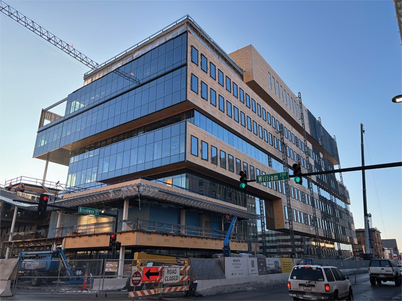 Modern glass building under construction at a city intersection. Cranes, fencing, and a "Road Closed" sign indicate active development work.