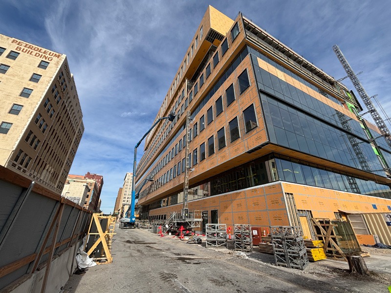 Construction site featuring a modern building with glass and orange sheathing, surrounded by scaffolding, against a bright blue sky. Tall building on the left.