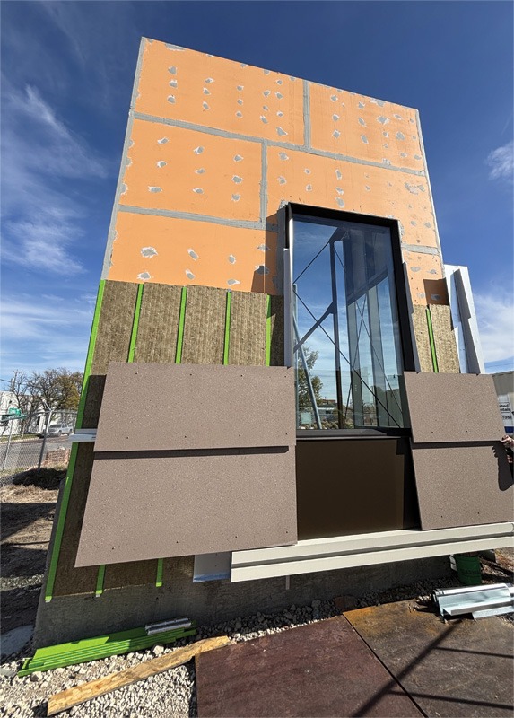A partially constructed building facade with orange sheathing, green insulation, and brown panels. A large window is centered, reflecting a clear sky.