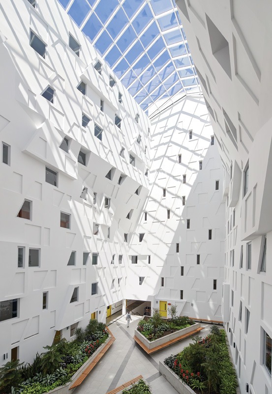 Modern atrium with a glass ceiling casting geometric shadows on white walls. Green plants and benches line the walkway, creating a bright, airy space.
