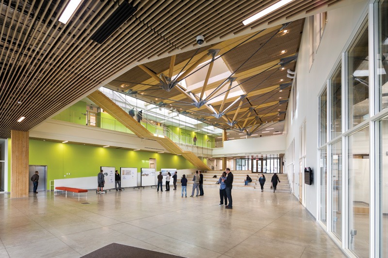 Modern interior with natural wood ceiling and large windows. People gather near green walls and an upstairs balcony, creating an open, airy atmosphere.