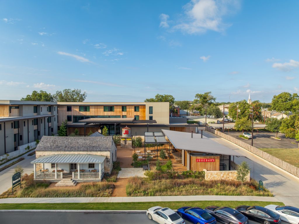 Aerial view of a modern complex featuring a rustic building with a metal roof, a patio area, and a contemporary structure with signage.