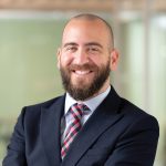 Smiling man with a beard in a suit and checkered tie, arms crossed. He stands in an office setting with a blurred background, conveying confidence.