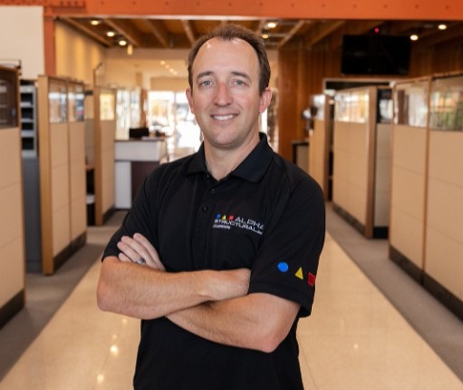 A man poses confidently in a modern office space, wearing a black polo shirt with colorful geometric shapes.