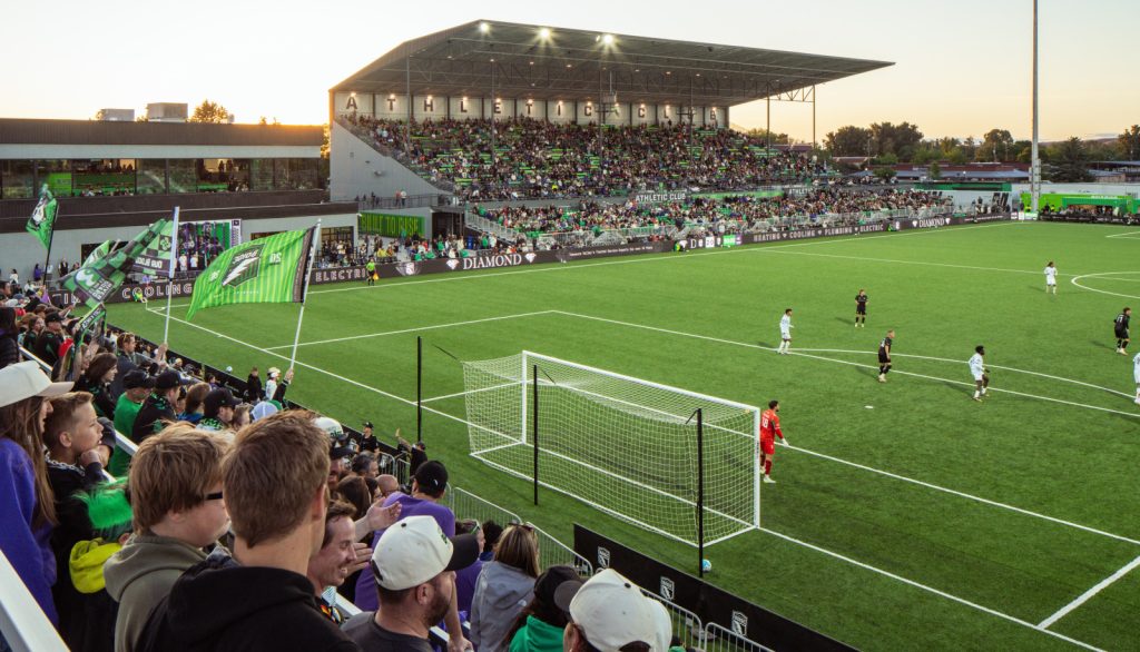 A view of a soccer match in a packed stadium with enthusiastic fans waving green flags. The field is bright green, and players are actively engaged in the game.