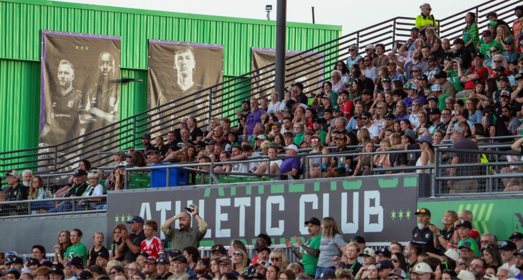 A crowded grandstand at a sports event with spectators of various ages, many wearing team jerseys, and large green banners displaying athletes in the background.