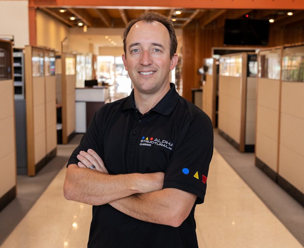 A man poses confidently in a modern office space, wearing a black polo shirt with colorful geometric shapes.