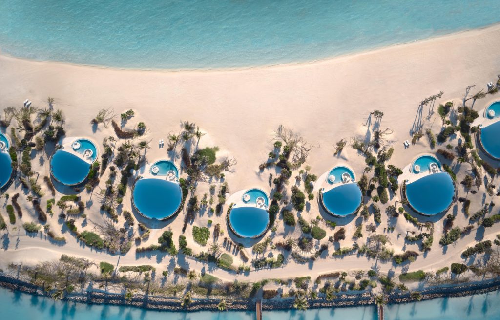 Aerial view of a beachfront resort featuring round swimming pools surrounded by palm trees, set against a light sandy beach and turquoise sea.