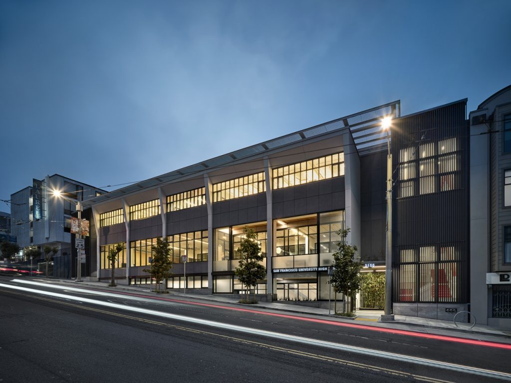 Modern building of San Francisco University High School at twilight, featuring large illuminated windows and architectural details, with light trails from passing vehicles.