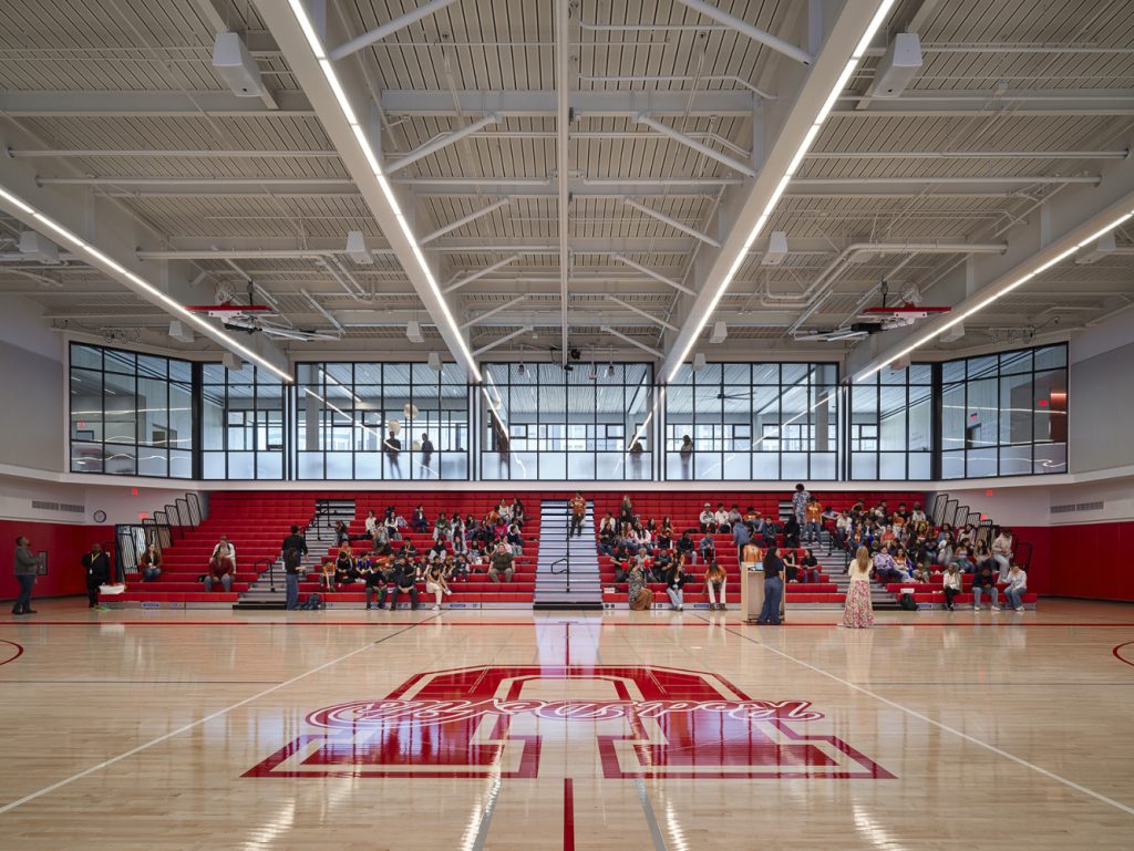 A spacious indoor basketball court with a polished wooden floor and red bleachers filled with spectators. Bright lights and large windows illuminate the space.