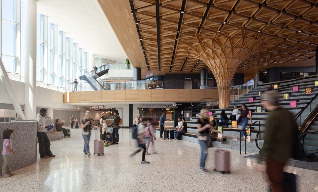 Spacious modern airport interior with wooden geometric ceiling and a unique tree-like column. People with luggage move briskly, conveying a dynamic ambiance.