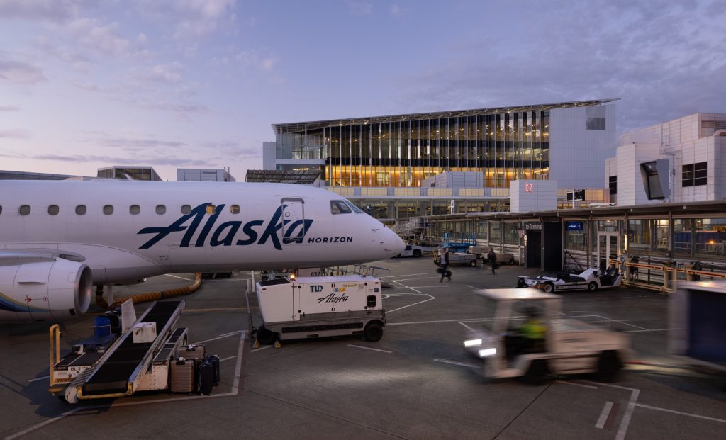 A parked Alaska Airlines plane at a bustling airport gate during twilight, with ground crew vehicles in motion and terminal lights glowing warmly.