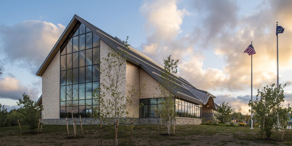Modern church with a steep roof and large windows reflecting a cloudy sunset sky. American flag flutters on a pole; trees dot the grassy yard.