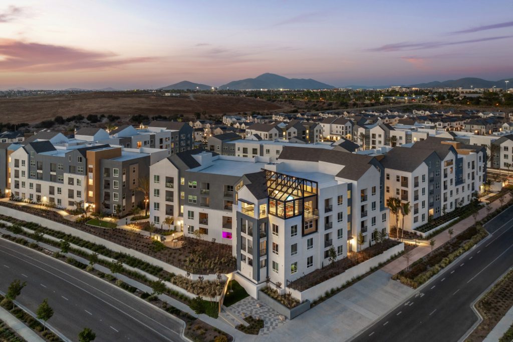 Modern residential complex with multi-story buildings, palm trees, and a sunset sky.
