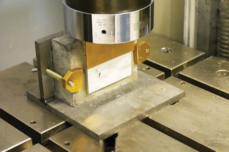 A metallic compression testing apparatus with a cylindrical top and a rectangular base holding a specimen, set against a backdrop of metallic plates.