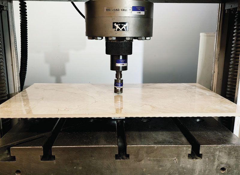 A mechanical press is positioned above a piece of glossy marble being tested for strength in a laboratory setting.