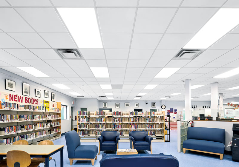 Interior of a library with bright white ceiling tiles, shelves filled with books, and blue armchairs around a wooden table.