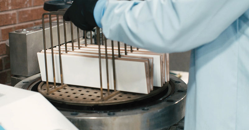 A person in a lab coat is holding a metal rack of rectangular white samples above a circular device on a work surface.