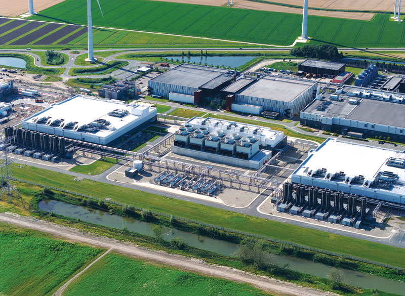 Aerial view of a data center complex surrounded by green fields and wind turbines, featuring multiple large buildings with flat roofs and visible cooling equipment.
