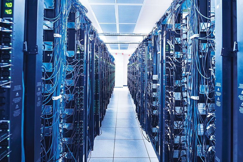 Interior of a data center with two rows of server racks, colorful LED indicators, and numerous hanging cables, illuminated by bright light at the end of the aisle.