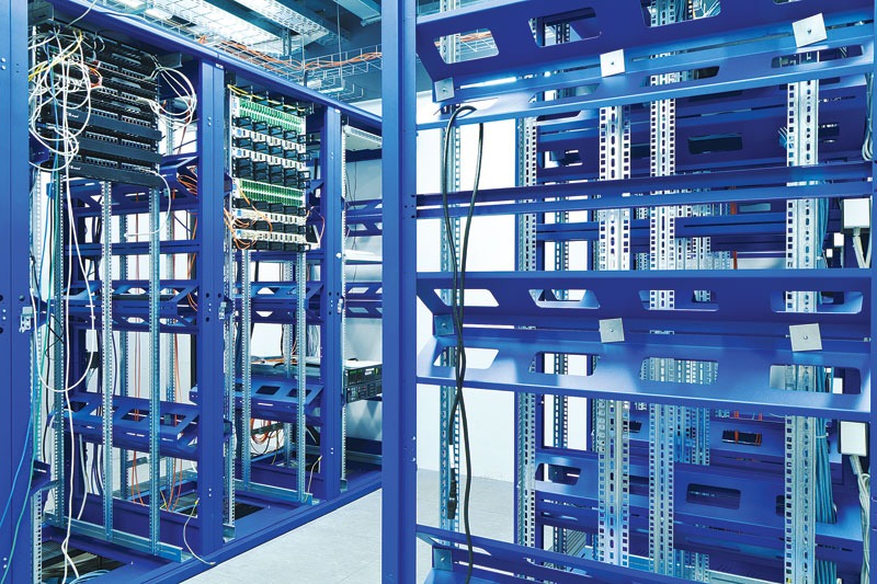 Interior view of a server room with blue metal racks filled with cables and networking equipment against a light wall.