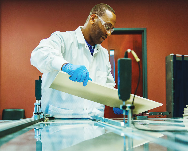 A man in a white lab coat and blue gloves is carefully lifting a beige sheet over a glass surface in a laboratory setting.