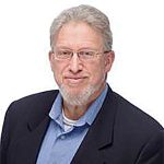 A middle-aged man with glasses and a light beard, wearing a dark blazer and blue shirt, smiles at the camera against a white background.