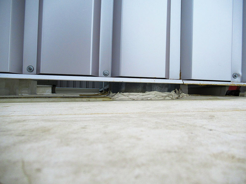 A low-angle view of a metallic storage unit showing a gap at the base, with grey panels and visible screws.