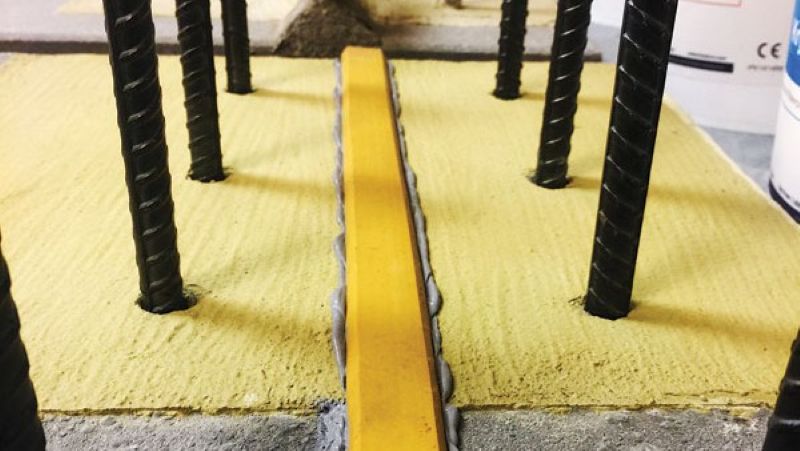 A close-up view of a construction setup featuring a yellow strip embedded in gray concrete, surrounded by vertical steel rebar rods.