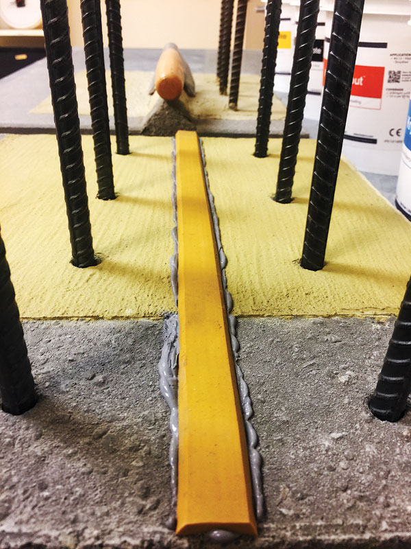 A close-up view of a construction setup featuring a yellow strip embedded in gray concrete, surrounded by vertical steel rebar rods.