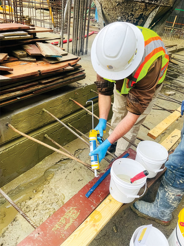 A construction worker in a hard hat and safety vest uses a caulking gun near buckets on a construction site with visible rebar and wooden planks.