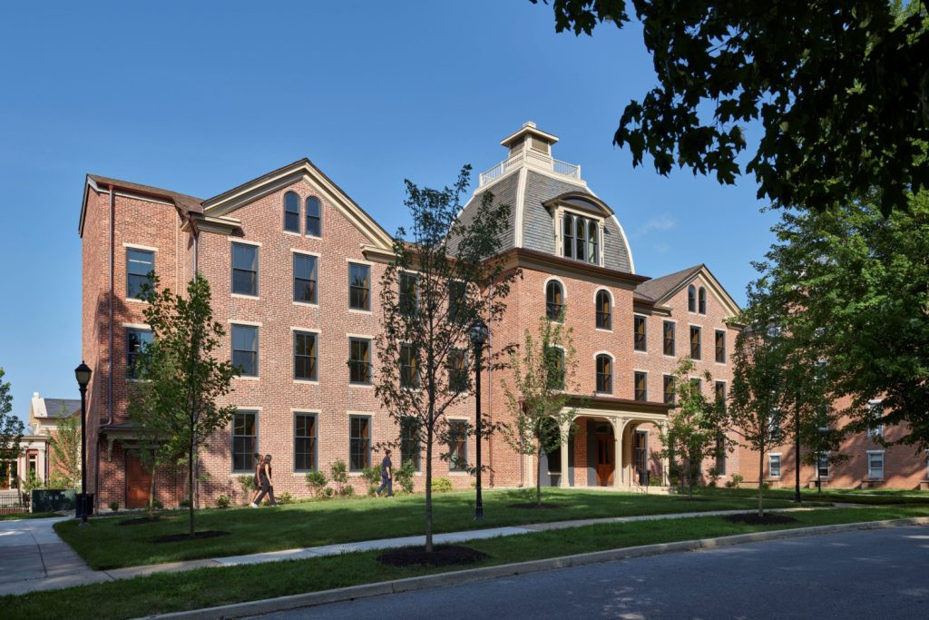 A red brick building featuring a central dome and gabled roof, with pedestrians walking along a landscaped sidewalk.