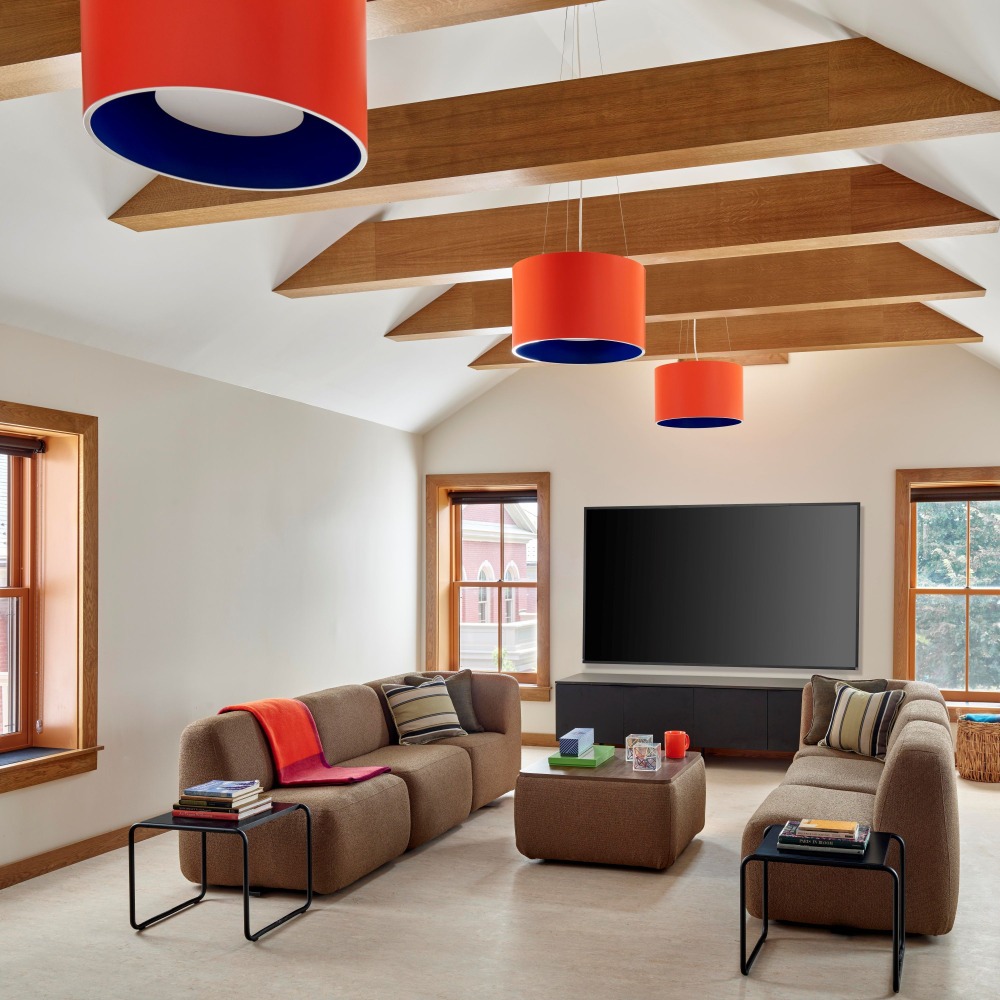 A bright living room with wooden beams, orange and blue pendant lights, two brown couches, a black television, and a square coffee table, filled with decorative objects.