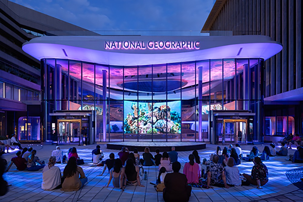 Exterior view of the National Geographic building at night, featuring vibrant lighting and a large screen showing underwater images, with people seated in front.