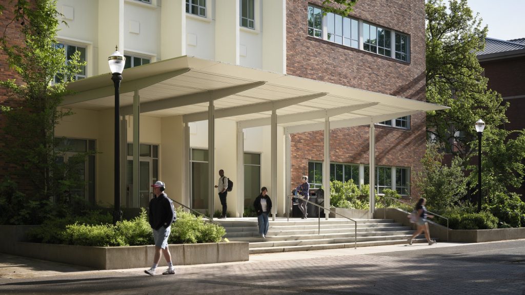 A modern academic building with a light-colored façade and brick wall, featuring a wide staircase, greenery, and people walking near the entrance.