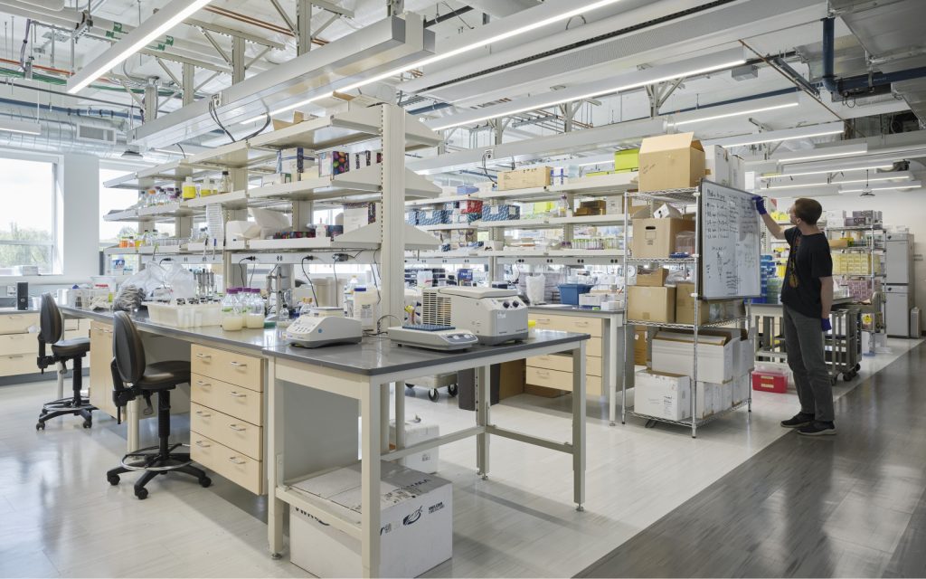 A modern laboratory with shelving stocked with colorful supplies and equipment, featuring a person writing on a whiteboard and various lab tables with scientific instruments.
