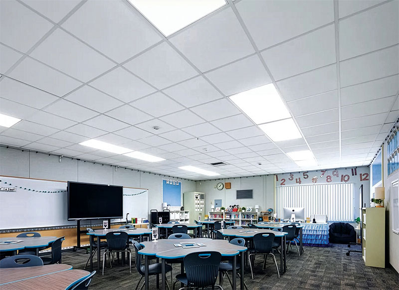 A bright classroom featuring circular tables and chairs, a digital screen on the wall, a whiteboard, and educational materials on shelves.