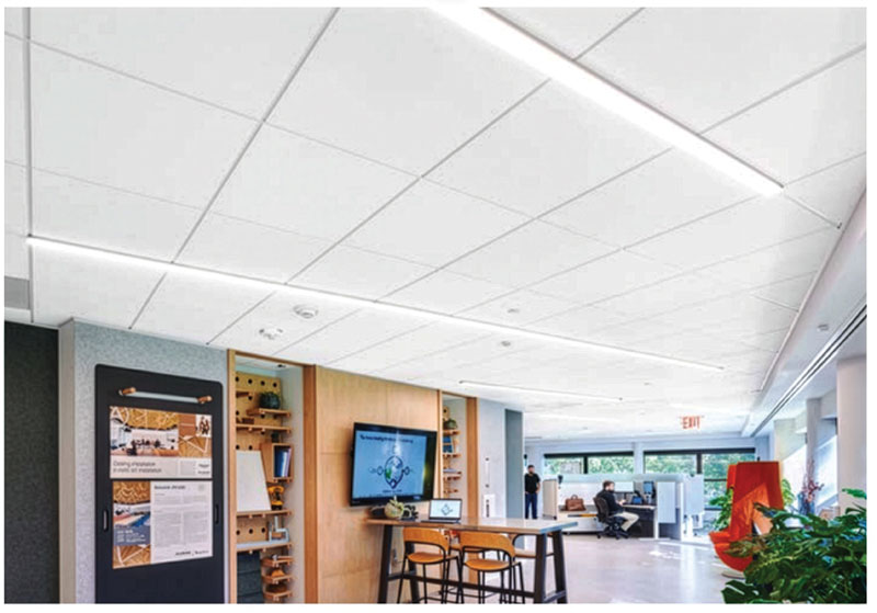 A modern office interior with linear ceiling lights, a bulletin board, a wooden table with chairs, and a wall-mounted screen displaying a molecular structure.