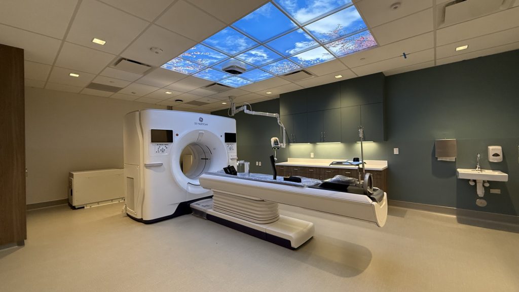 A modern medical room with a CT scanner, examination table, and a ceiling that mimics a blue sky with cherry blossoms, creating a calming atmosphere.