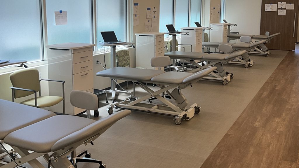 A health care facility with several adjustable treatment tables, light green chairs, and white storage units, all arranged in a bright, organized room.