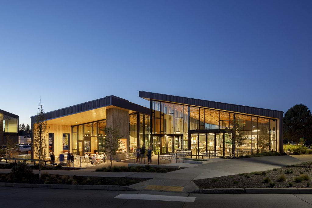 A modern building at dusk with large glass panels, wooden and metal elements, surrounded by landscaped pathways and trees.
