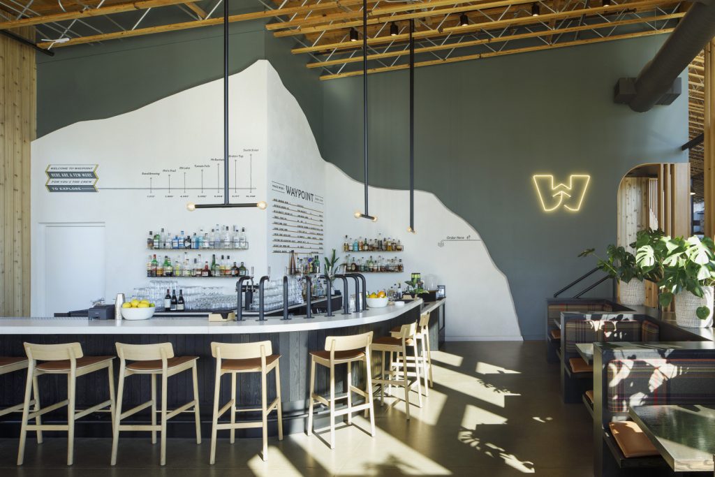 Interior view of a modern bar named "Waypoint" featuring a curved white bar, liquor shelves, a mountain mural on the wall, and wooden stools. A neon "W" sign is visible, along with cozy booths in the background.