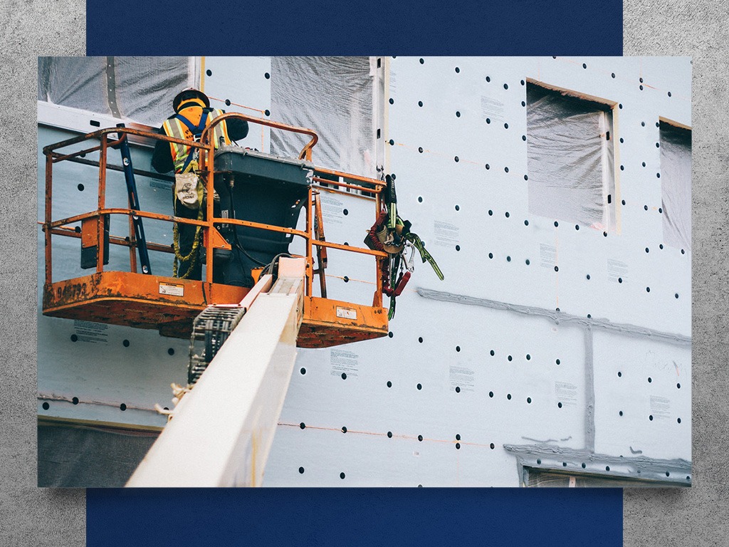 A construction worker in a boom lift, wearing a yellow vest and hard hat, stands before a wall with multiple holes during construction.