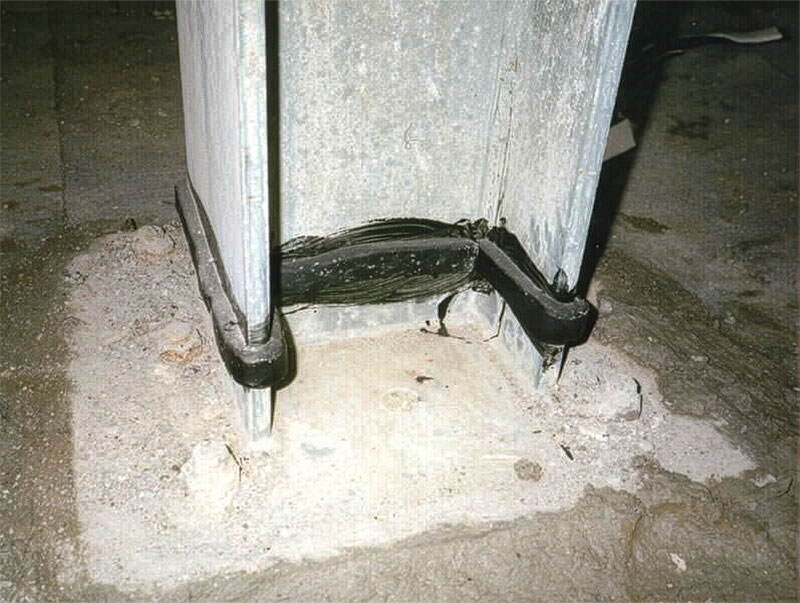Base of a weathered metal column secured to a concrete floor with black brackets, showing signs of wear and rust.